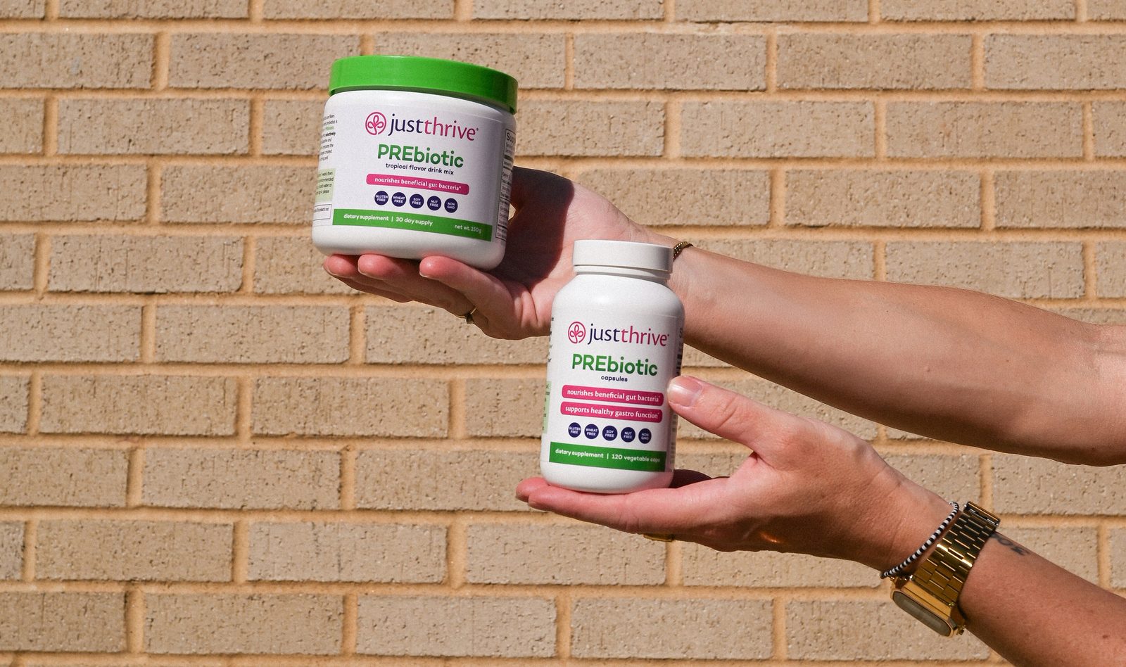 Two hands holding Just Thrive PREbiotic drink mix and capsules against a brick wall