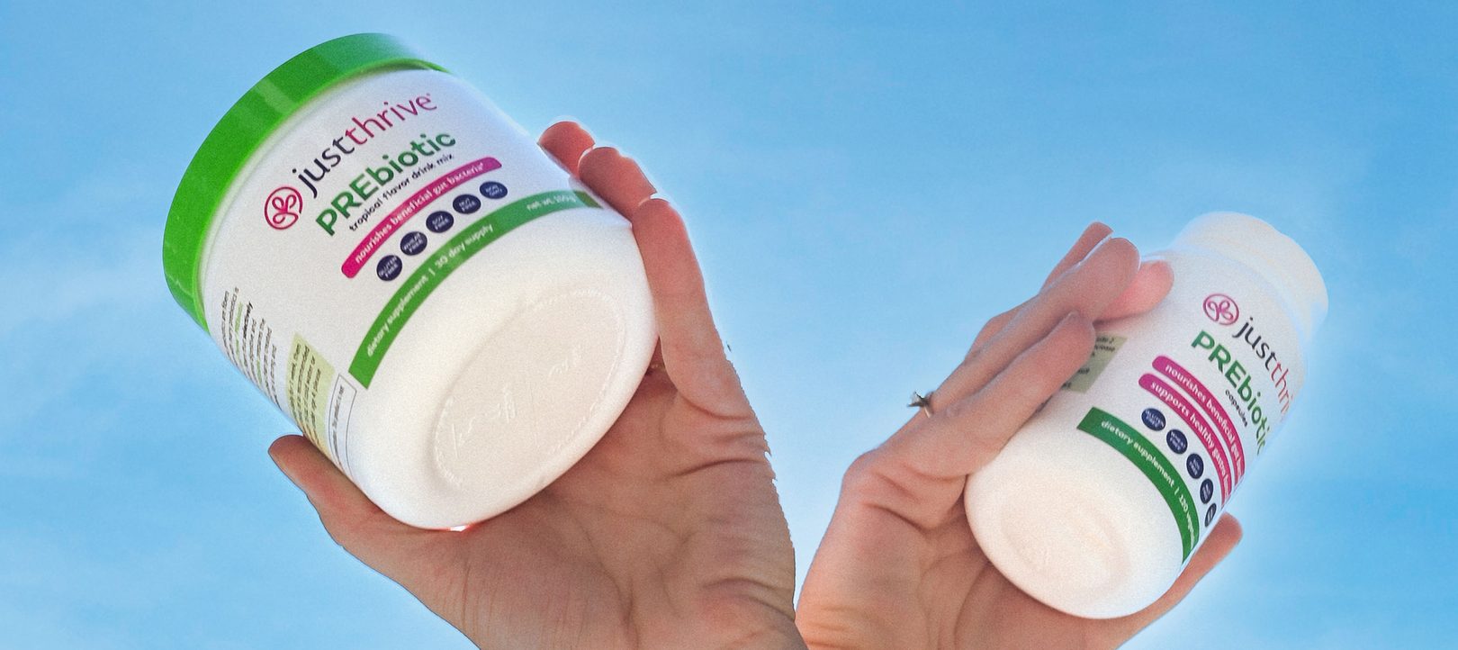 Two hands holding Just Thrive PREbiotic containers against a blue sky