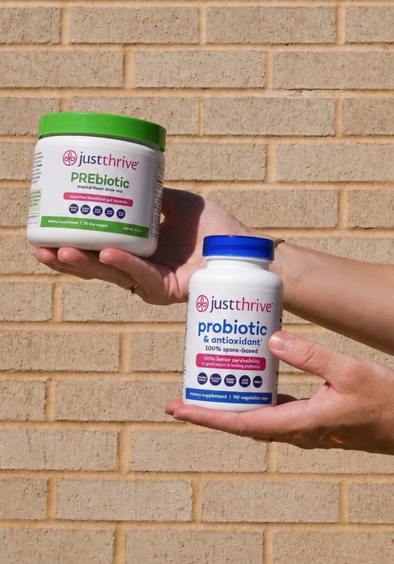 Two hands holding Just Thrive PREbiotic and Just Thrive Probiotic against a brick wall